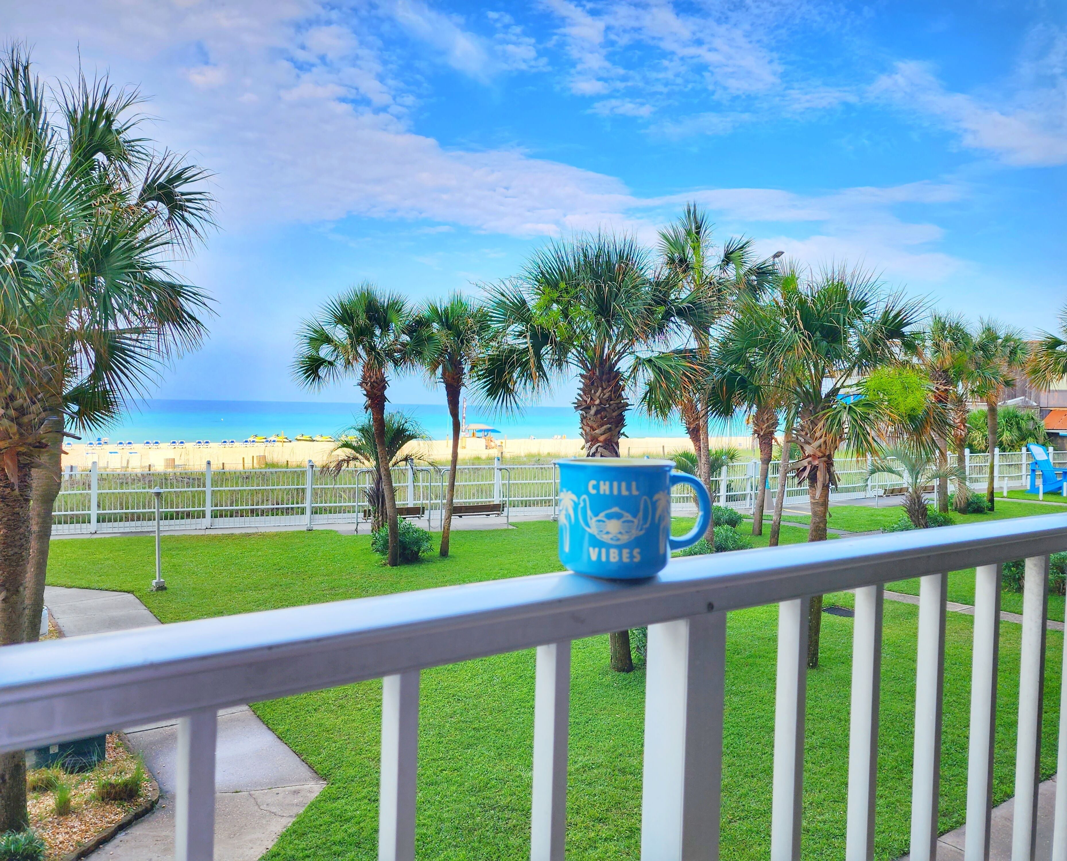 A balcony with a railing, a blue mug with "CHILL VIBES" text, palm trees, and a view of the beach.