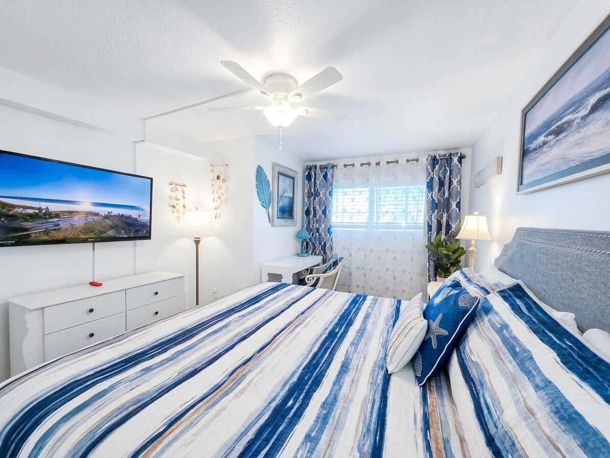 Une chambre à coucher comprenant un lit, un bureau, une chaise, un ventilateur de plafond, une télévision et une fenêtre avec des rideaux.