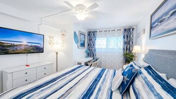 Une chambre à coucher comprenant un lit, un bureau, une chaise, un ventilateur de plafond, une télévision et une fenêtre avec des rideaux.