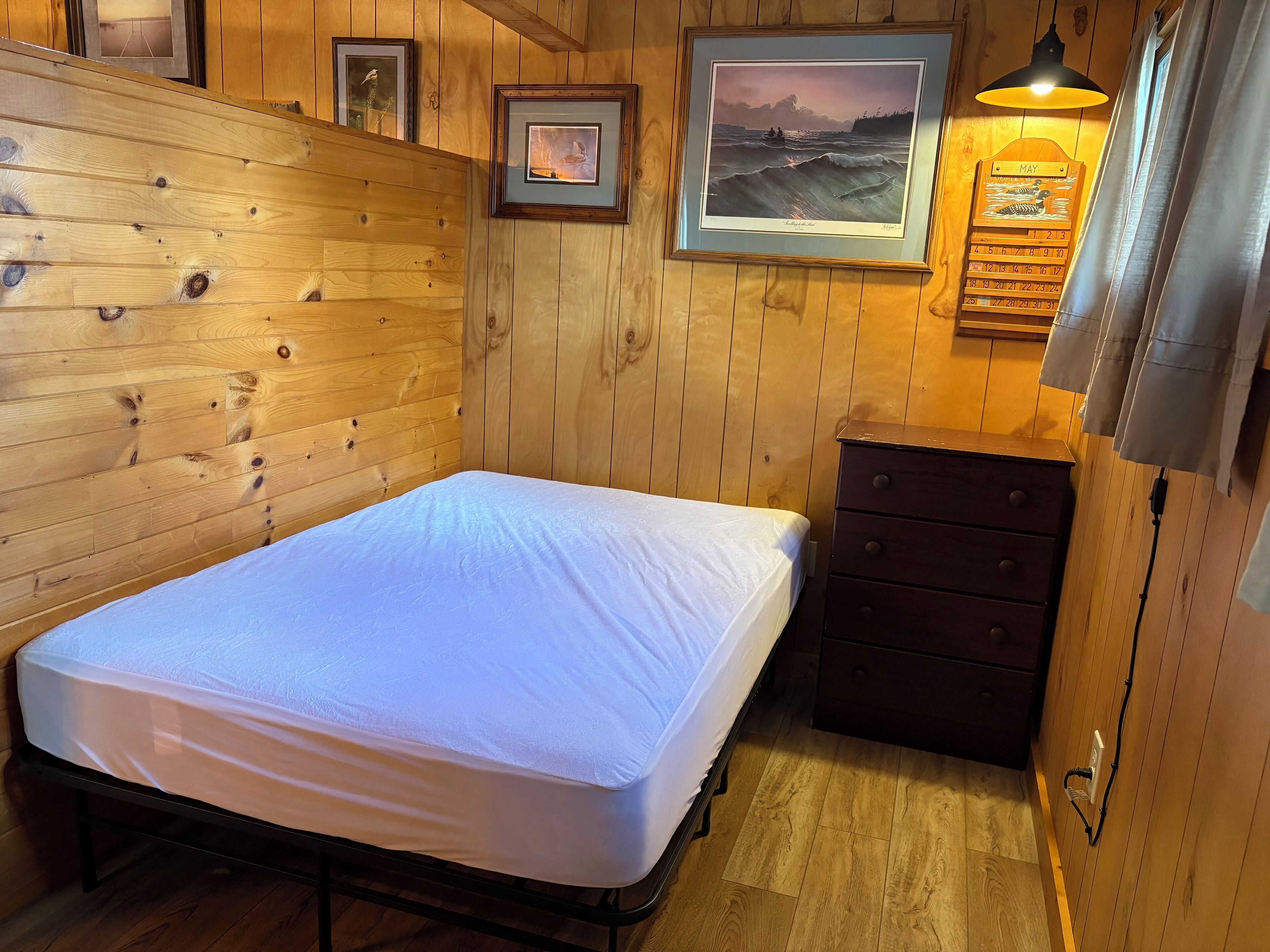 A wooden-paneled room with a bed, a dresser, framed pictures, and a wall-mounted calendar.