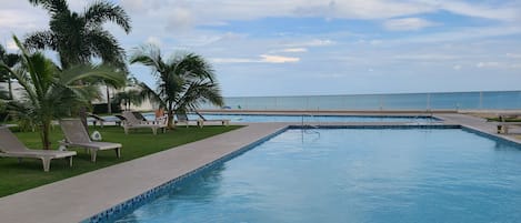 Una piscina con sillas de descanso y palmeras, rodeada de un cielo despejado.