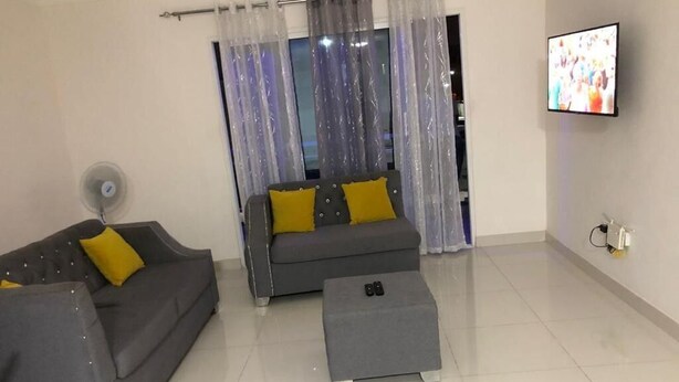 Un salon avec deux canapés gris, dont l’un est agrémenté de coussins jaunes, un pouf gris, un téléviseur fixé au mur et une fenêtre ornée de rideaux légers.