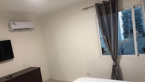 Une chambre à coucher comprenant un lit, une commode en bois foncé, un téléviseur à écran plat, un climatiseur et une fenêtre avec des rideaux.