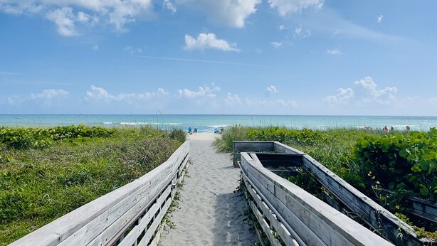 Ein hölzerner Steg führt zu einem Strand mit blauem Himmel und ruhigem Wasser.