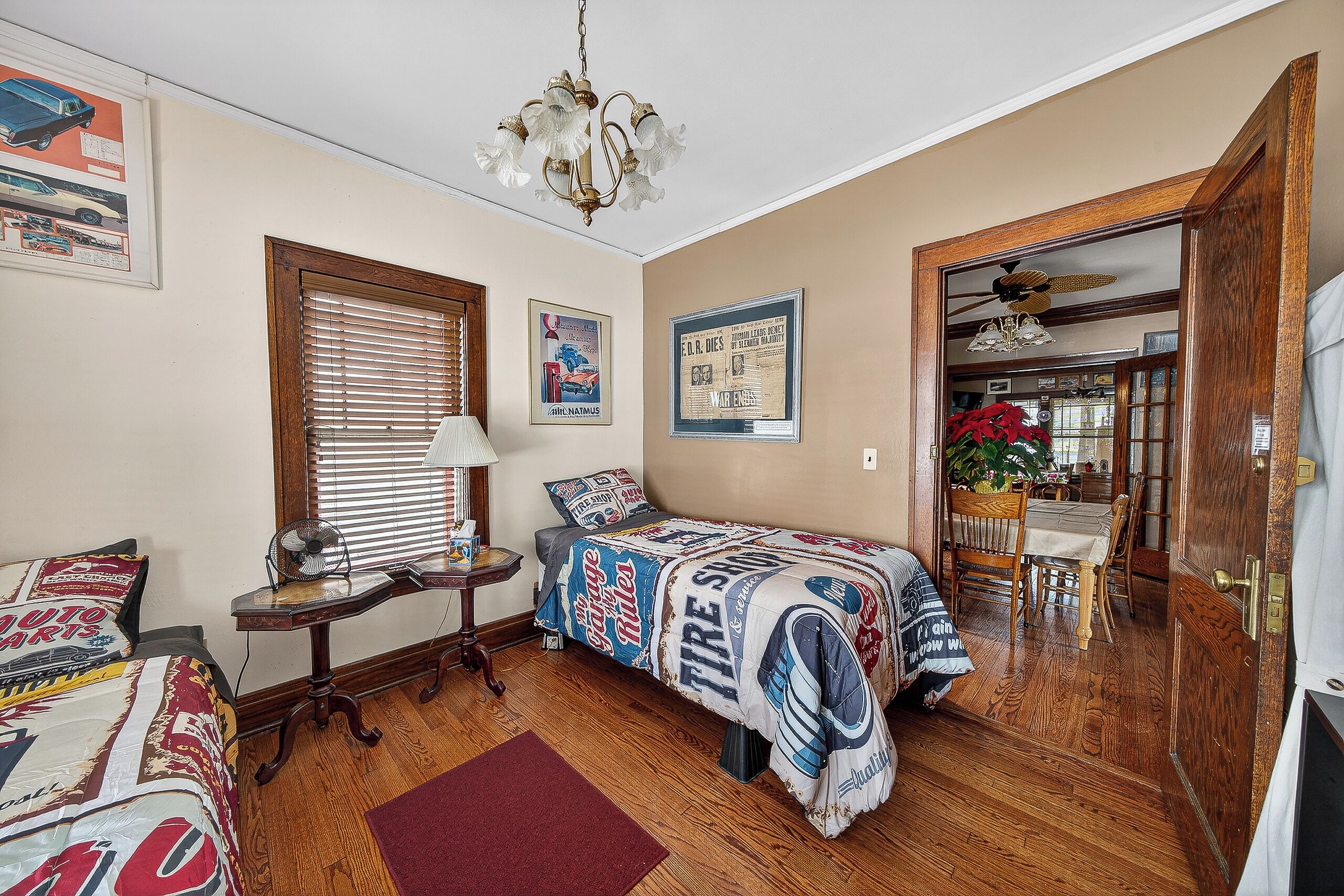 A bedroom with two beds, a wooden floor, a ceiling fan, and a dining area visible through an open door.