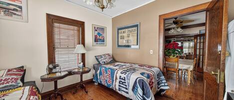 A bedroom with two beds, a wooden floor, a ceiling fan, and a dining area visible through an open door.