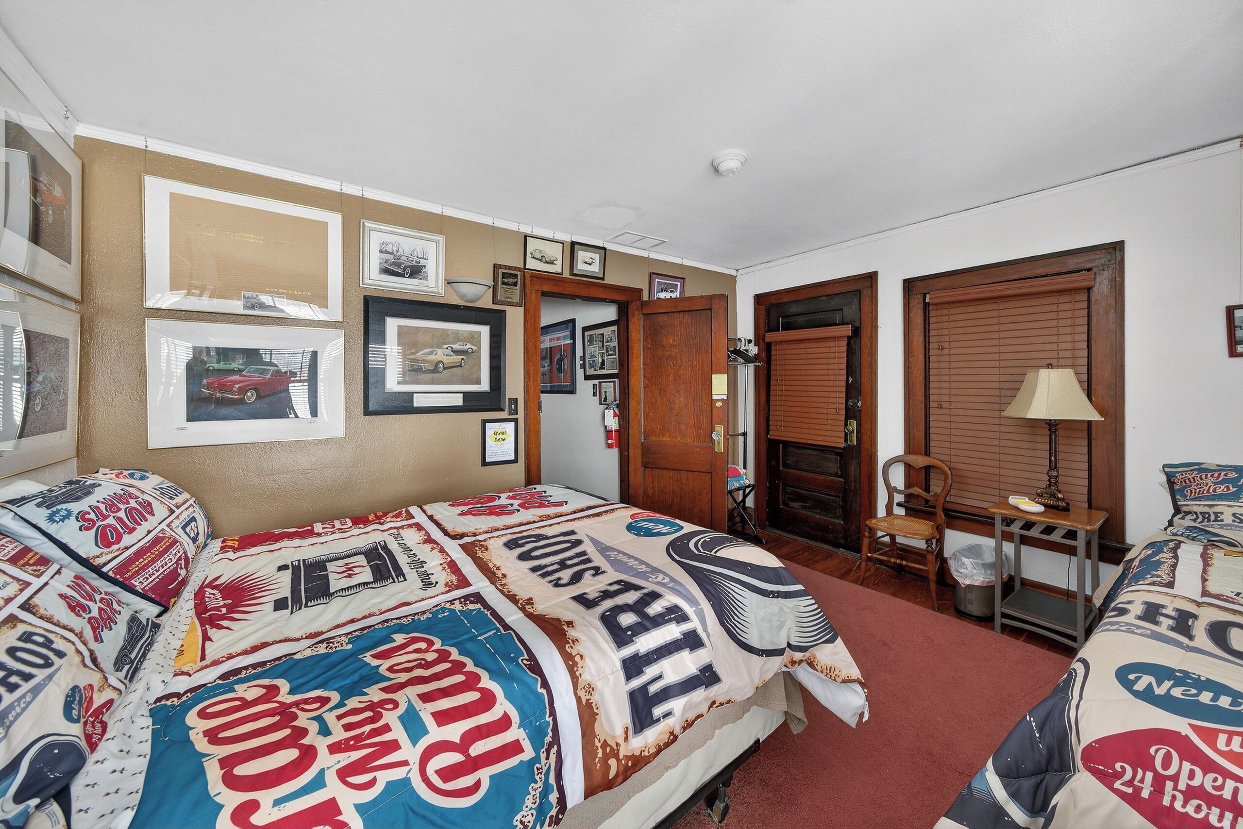 Een hotelkamer met een groot bed belegd met een plaid met een vintage thema, een houten stoel, een klein tafeltje met een lamp en lijstjes met foto’s aan de muur.