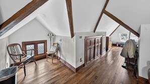 A spacious attic room with wooden beams, a large window, and a cozy seating area with a chair and table. - The Avanti Houses (South Bend)