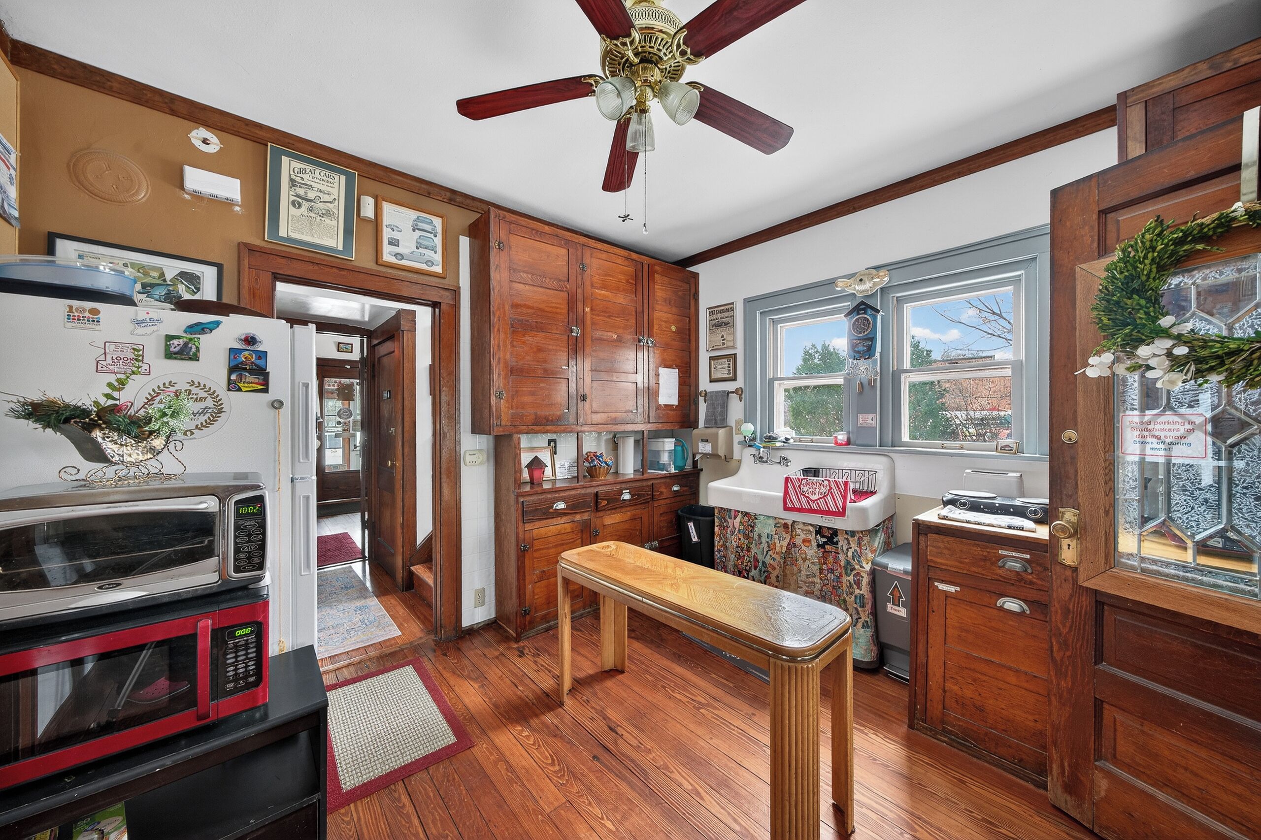 Una cocina con armarios de madera, ventilador de techo, refrigerador, microondas, una mesa de madera y una ventana con vista a árboles.