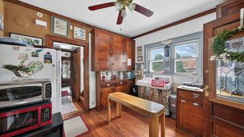 Una cocina con armarios de madera, ventilador de techo, refrigerador, microondas, una mesa de madera y una ventana con vista a árboles.