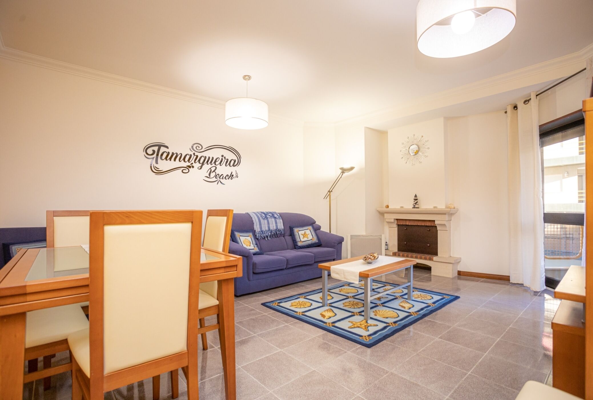 A living room with a sofa, a dining table with chairs, a fireplace, and a wall with a sign that reads "Tamariguera Beach".