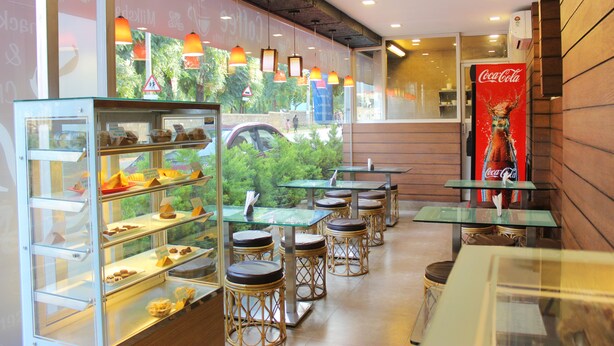 A café with glass display cases of pastries, wicker stools, and tables. - Panchvati Comforts (Bengaluru)