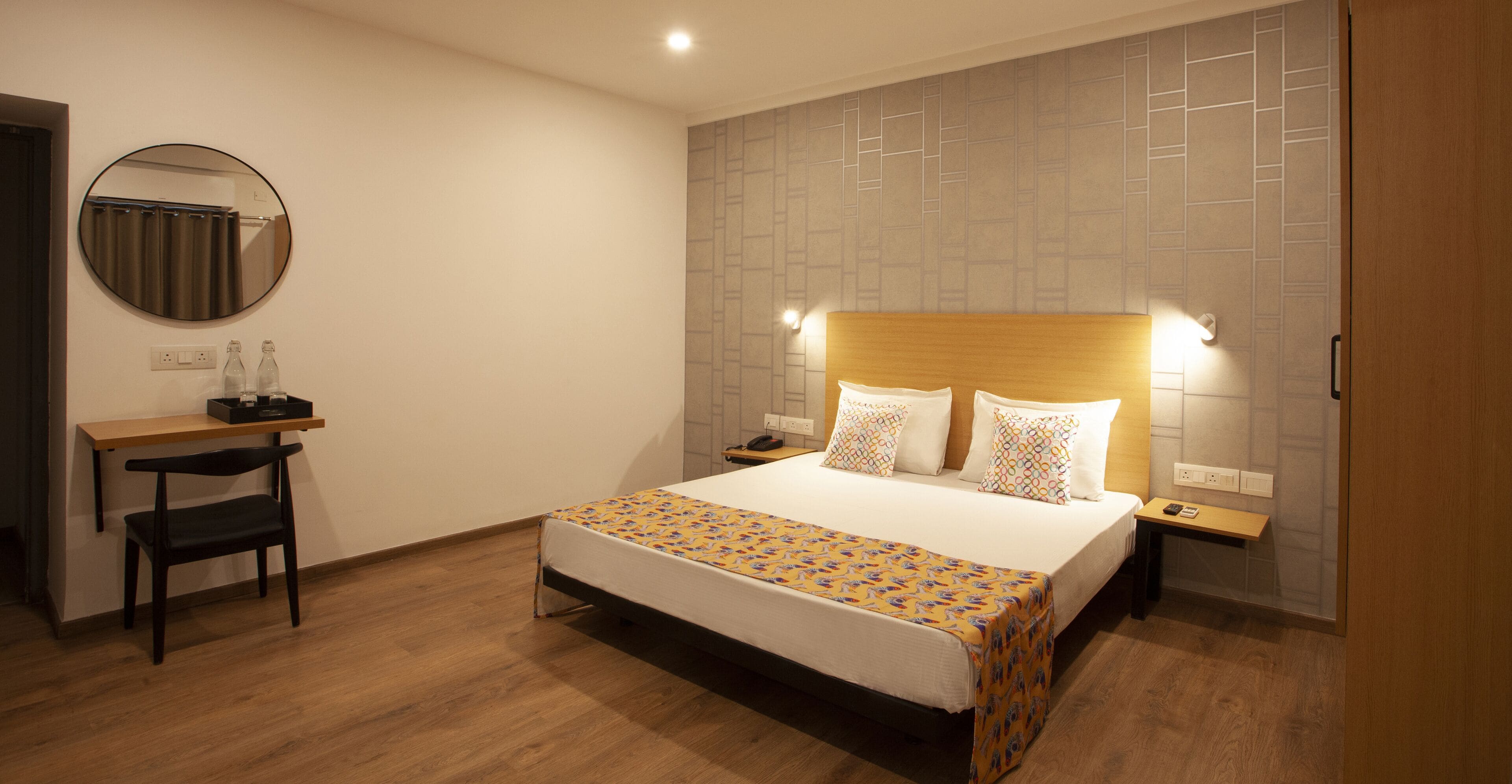 A hotel room with a bed, bedside tables, a desk with a chair, a round mirror, and a wall with a geometric pattern.