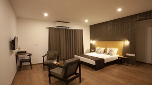 A modern hotel room with a wooden floor, a bed with a headboard, a desk with a chair, a TV mounted on the wall, and a curtain. - Panchvati Comforts (Bengaluru)