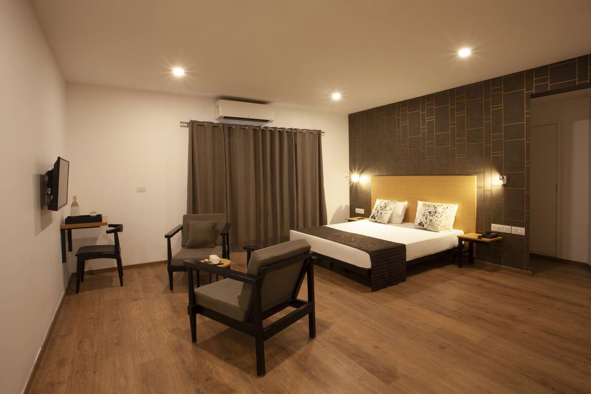 Une chambre d’hôtel moderne avec un plancher en bois, un lit doté d’une tête de lit, un bureau avec une chaise, un téléviseur fixé au mur et un rideau.