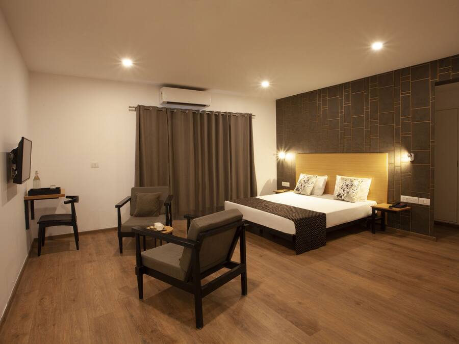 A modern hotel room with a wooden floor, a bed with a headboard, a desk with a chair, a TV mounted on the wall, and a curtain.