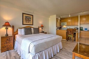 A bedroom with a large bed, two bedside tables with lamps, a wooden nightstand, a framed picture on the wall, and a kitchenette with wooden cabinets and a microwave.