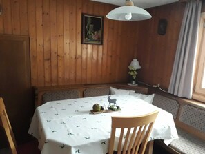 Zona pranzo con tavolo rotondo ricoperto da una tovaglia bianca a fantasia, sedie in legno, una panca, una lampada a sospensione e un quadro appeso al muro.
