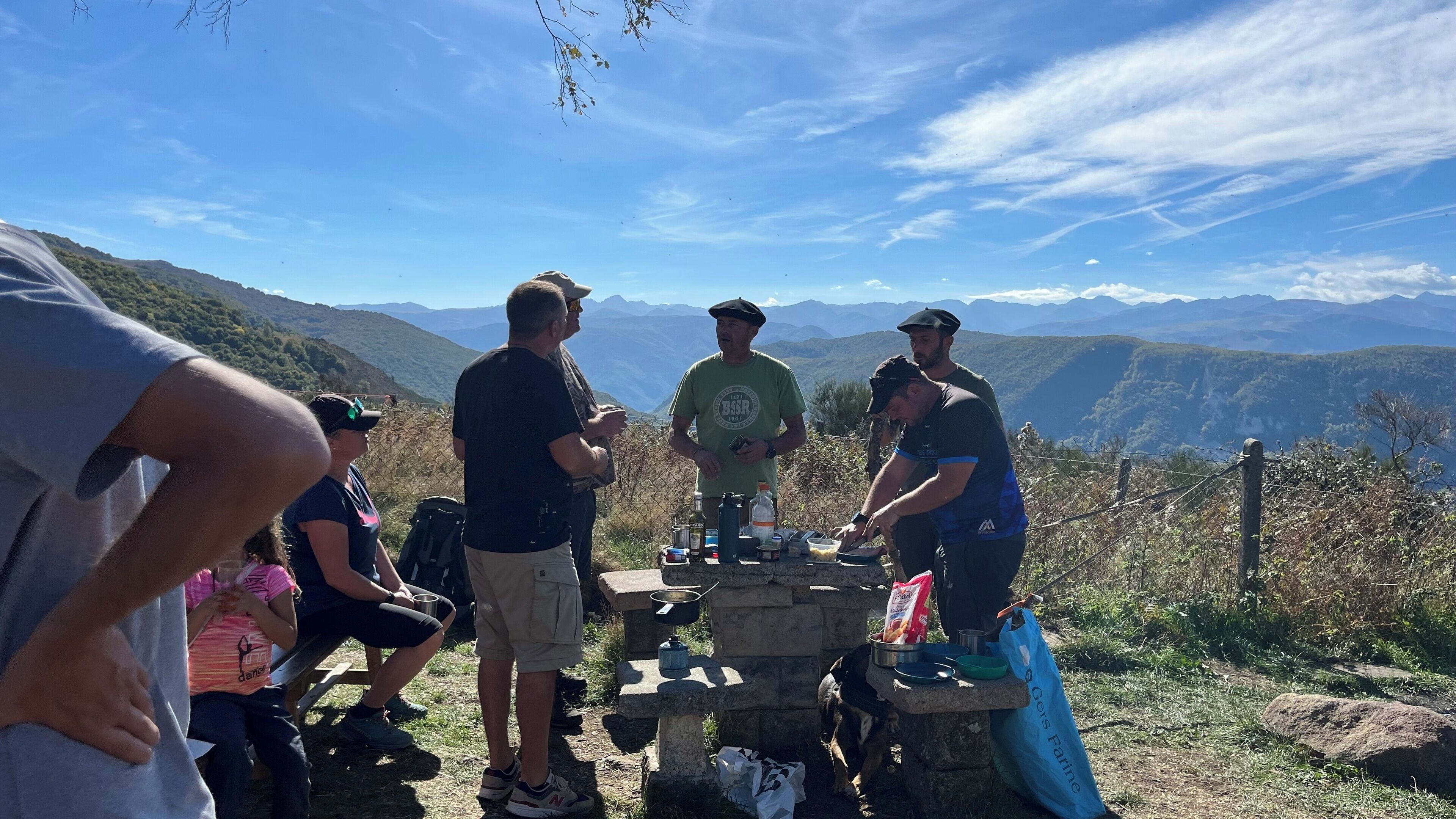 Een groep mensen verzameld rond een stenen tafel met verschillende soorten eten en een hond, met uitzicht op een bergachtig landschap.