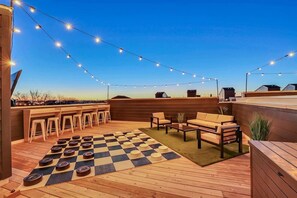 A rooftop deck with wooden flooring, a checkered mat, outdoor seating, and string lights.