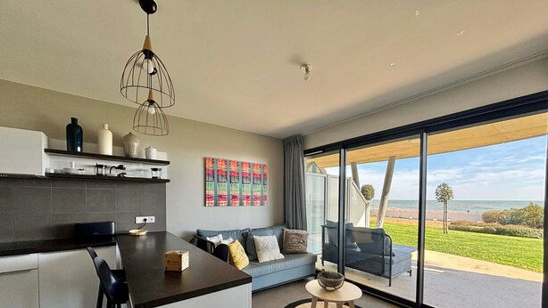 A modern kitchen with a central island, a dining area with a sofa and chairs, and a large sliding glass door leading to a patio with a view of the ocean.