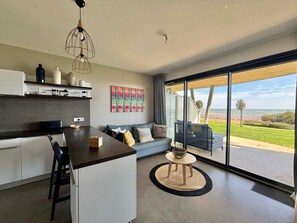 A modern kitchen with a central island, a dining area with a sofa and chairs, and a large sliding glass door leading to a patio with a view of the ocean. - Seafront apartment, 2 bedrooms, pool, parking, pets allowed (La Tranche-sur-Mer)
