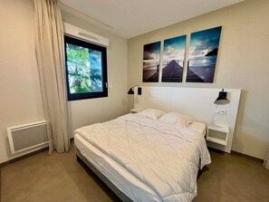 Ein Schlafzimmer mit einem Bett, einem Fenster mit Vorhängen, einem Nachttisch, einer Lampe und drei an der Wand hängenden Bildern.