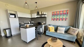 A modern kitchen with white cabinets, a central island, and pendant lighting. A blue sofa with decorative pillows is in the living area, and a small round table with a woven rug is in the foreground.