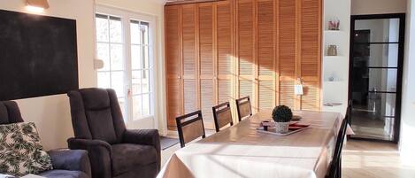 A dining area with a long table, chairs, a hanging light fixture, and a wooden wall with shutters.