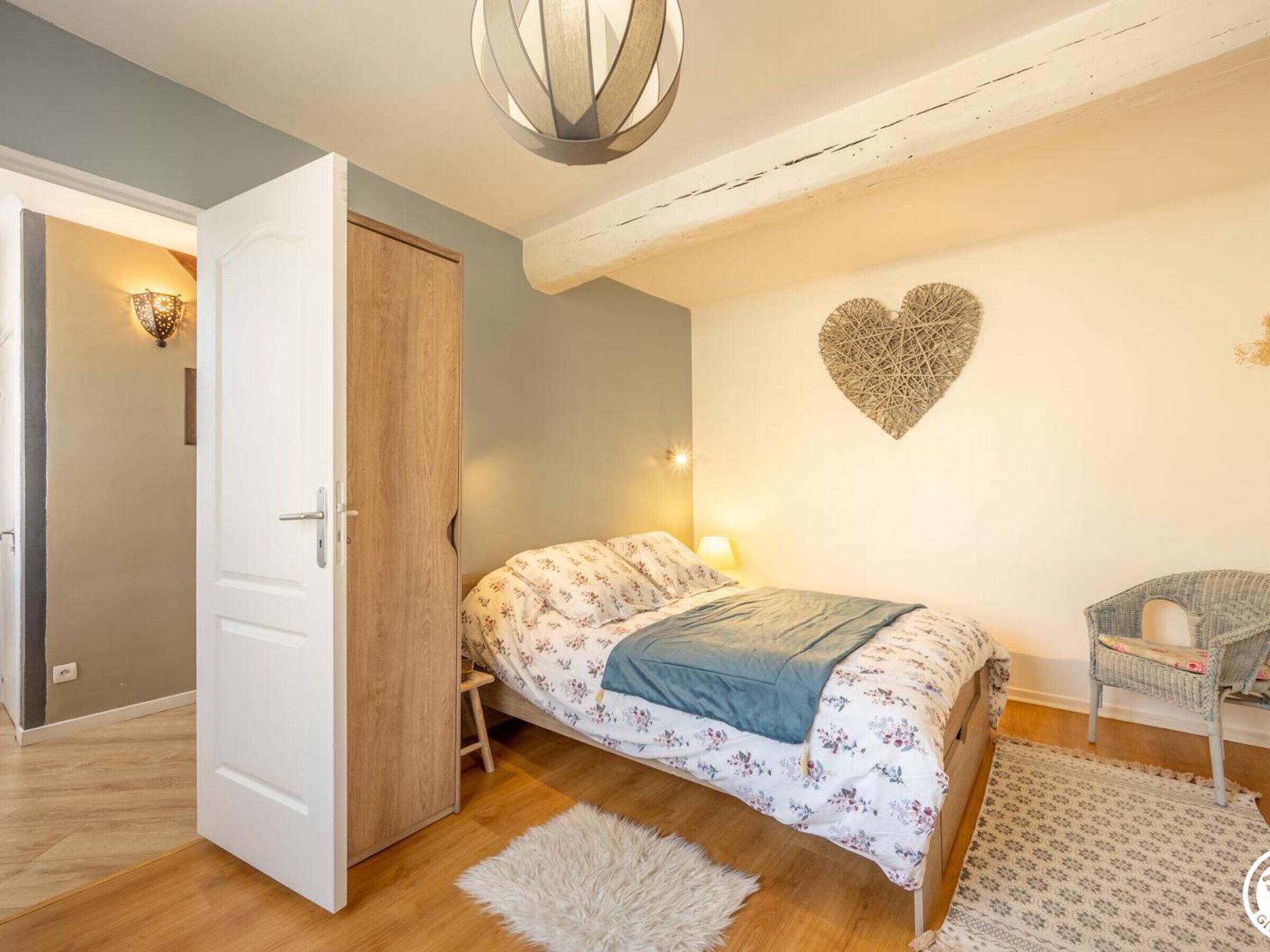 A bedroom with a bed, a chair, a heart-shaped wall decoration, and a door leading to another room.