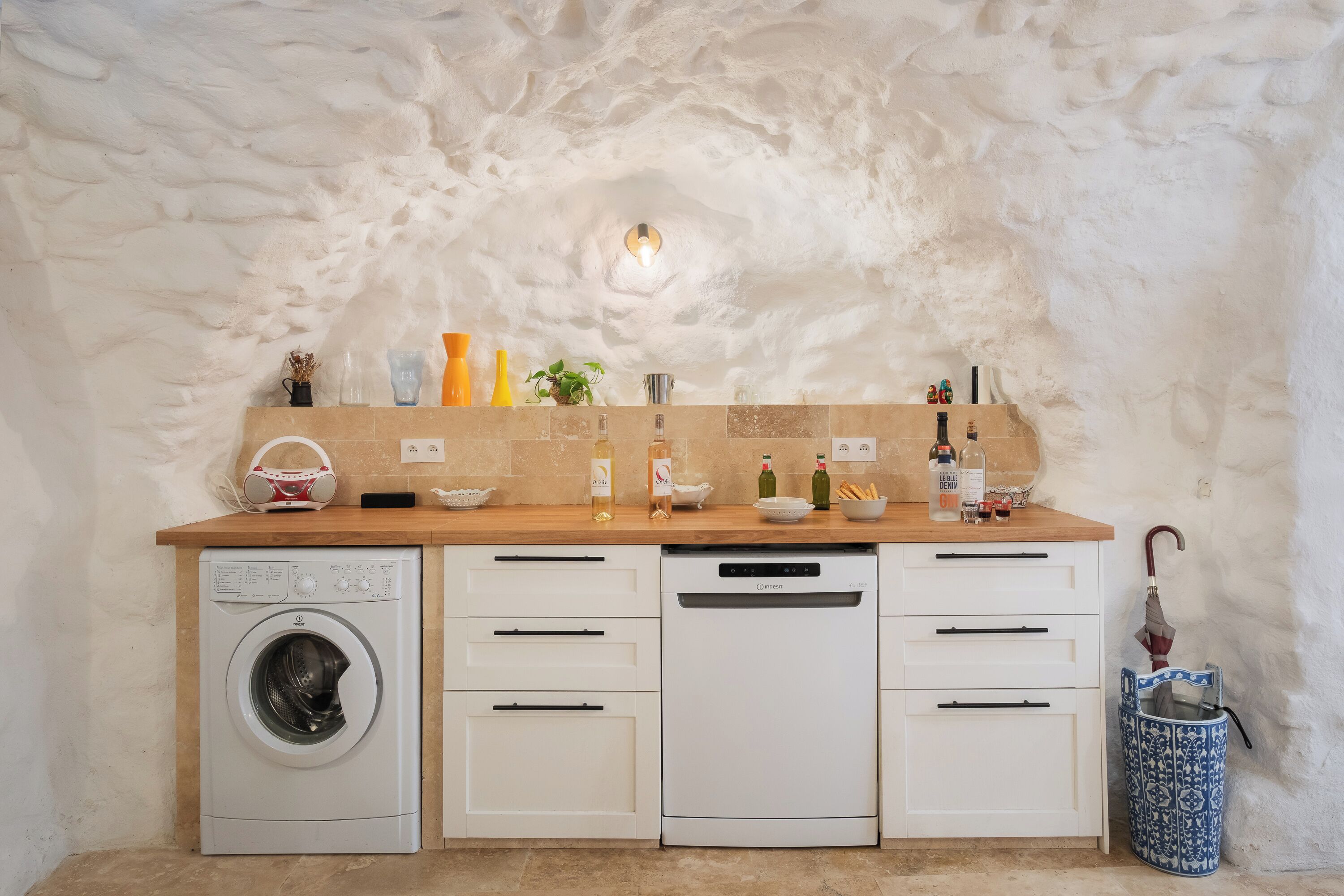 Une cuisine compacte équipée d’un lave-linge, d’un lave-vaisselle et d’un plan de travail en bois sur lequel sont disposés divers objets.