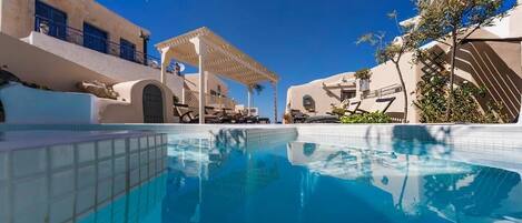 Ein Poolbereich mit klarem, blauem Pool, weißen Gebäuden und einer Pergola.