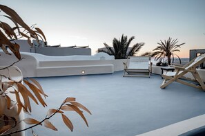 A rooftop terrace with lounge chairs, palm trees, and a clear sky. - Lotza Studios (Santorini)