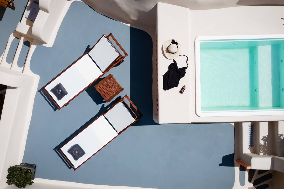 A top-down view of a sunlit pool area with two lounge chairs, a small table, and a bottle of sunscreen.
