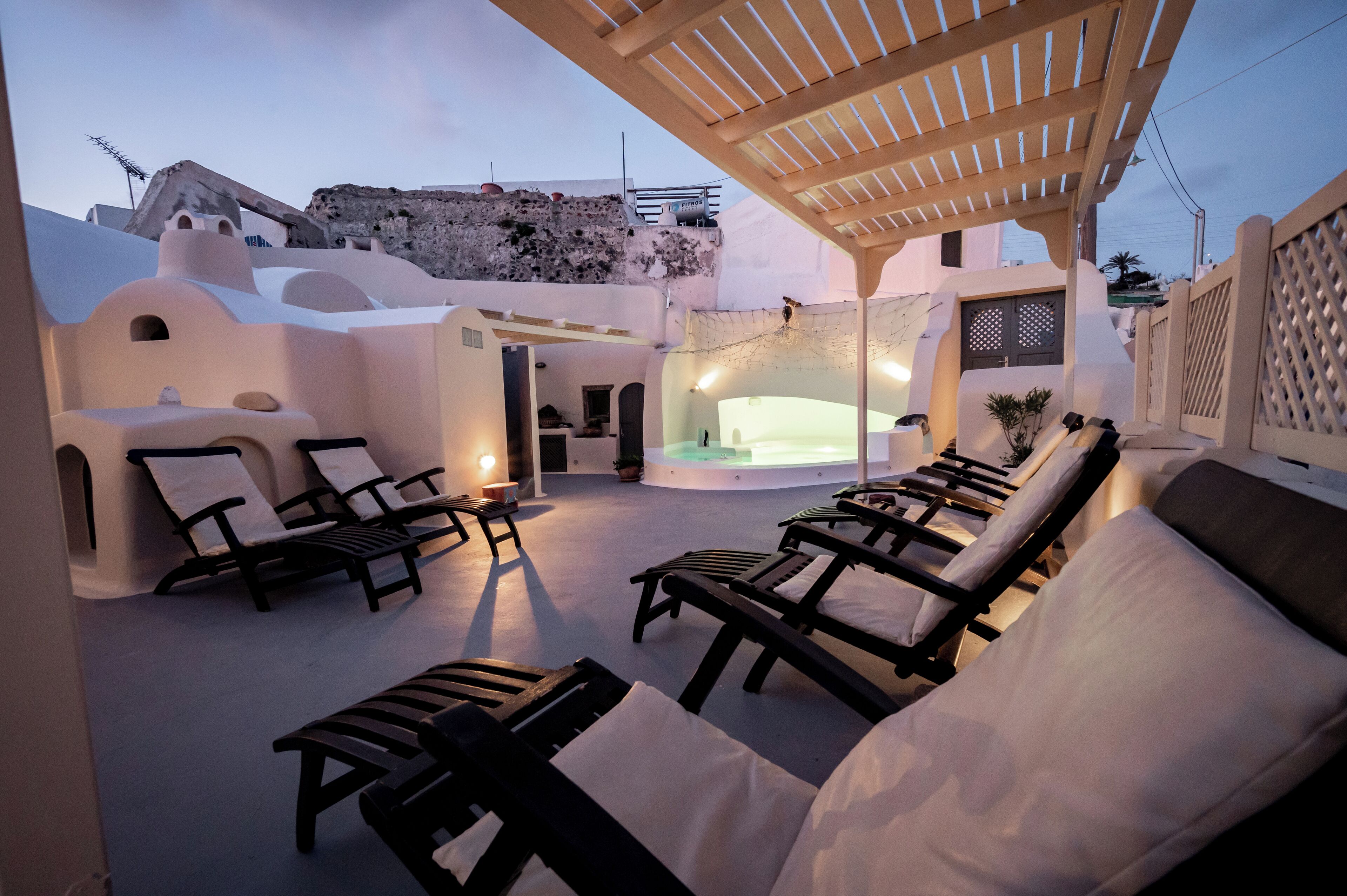 A sunlit outdoor seating area with lounge chairs and a hot tub, surrounded by white-washed buildings with unique architecture.