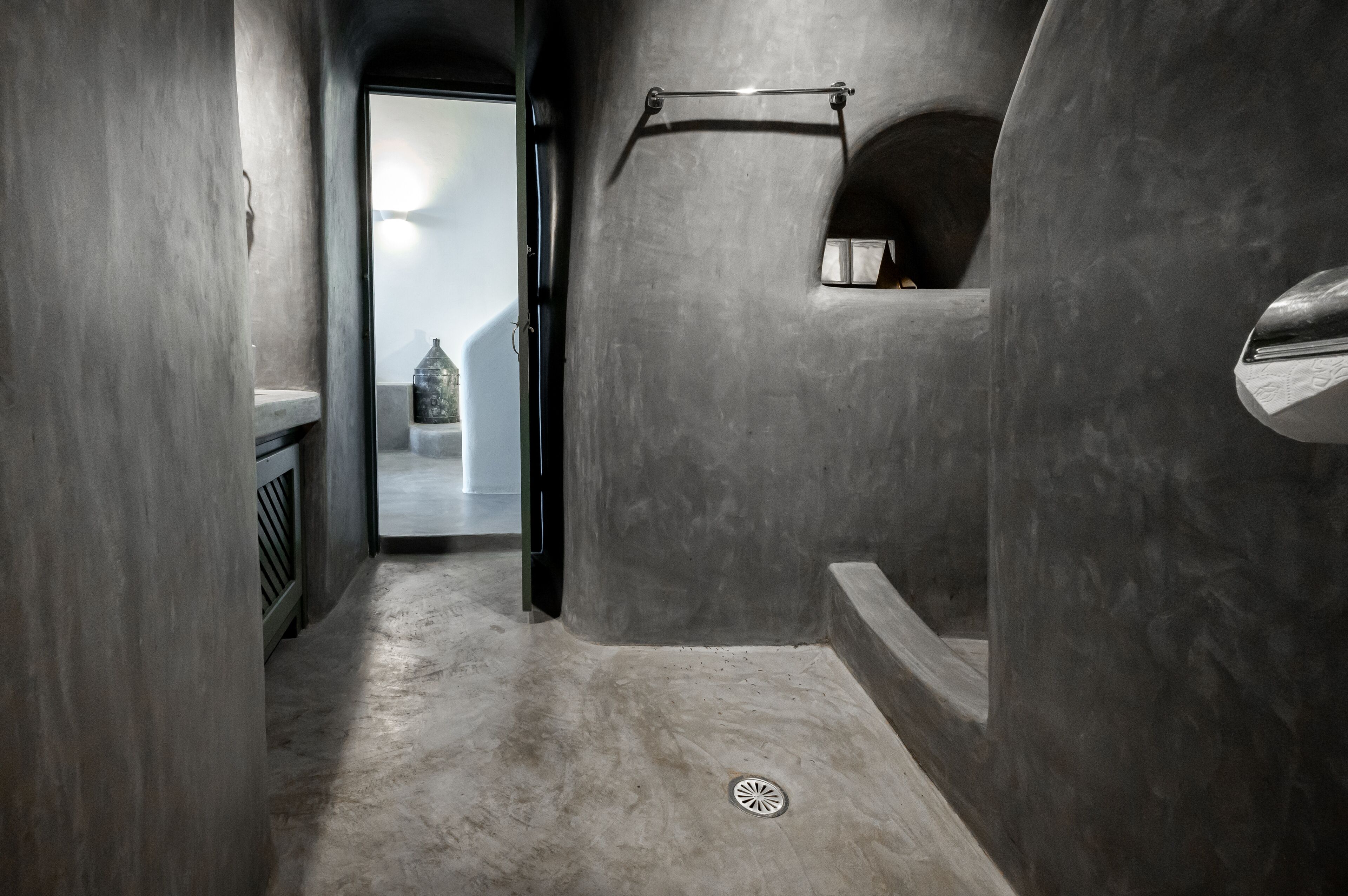 A narrow, dimly lit hallway with a concrete floor and walls, leading to a door with a view of a building outside.