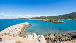 A coastal view with a rocky breakwater, clear blue water, and distant hills.