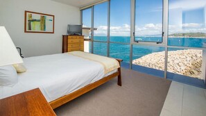 A bedroom with a bed, a television on a wooden dresser, and a large window with a view of the sea and rocky beach.