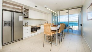 A modern kitchen with a built-in refrigerator, oven, and microwave. There is a dining area with wicker chairs and a table. The room has a large sliding glass door leading to a balcony with a view of the sea. - 1 Bright Point Apartment 3303 (Nelly Bay)