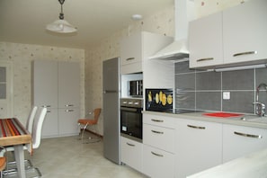 Eine moderne Küche mit weißen Schränken, einem Edelstahl-Kühlschrank, einem Backofen und einem Spülbecken. Ein Esstisch mit Stühlen und ein Stuhl mit rotem Sitzkissen sind ebenfalls vorhanden.