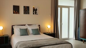 Un letto rifatto con lenzuola bianche e due cuscini, affiancato da comodini con lampade, in una stanza con un'ampia finestra e vista sull'esterno.
