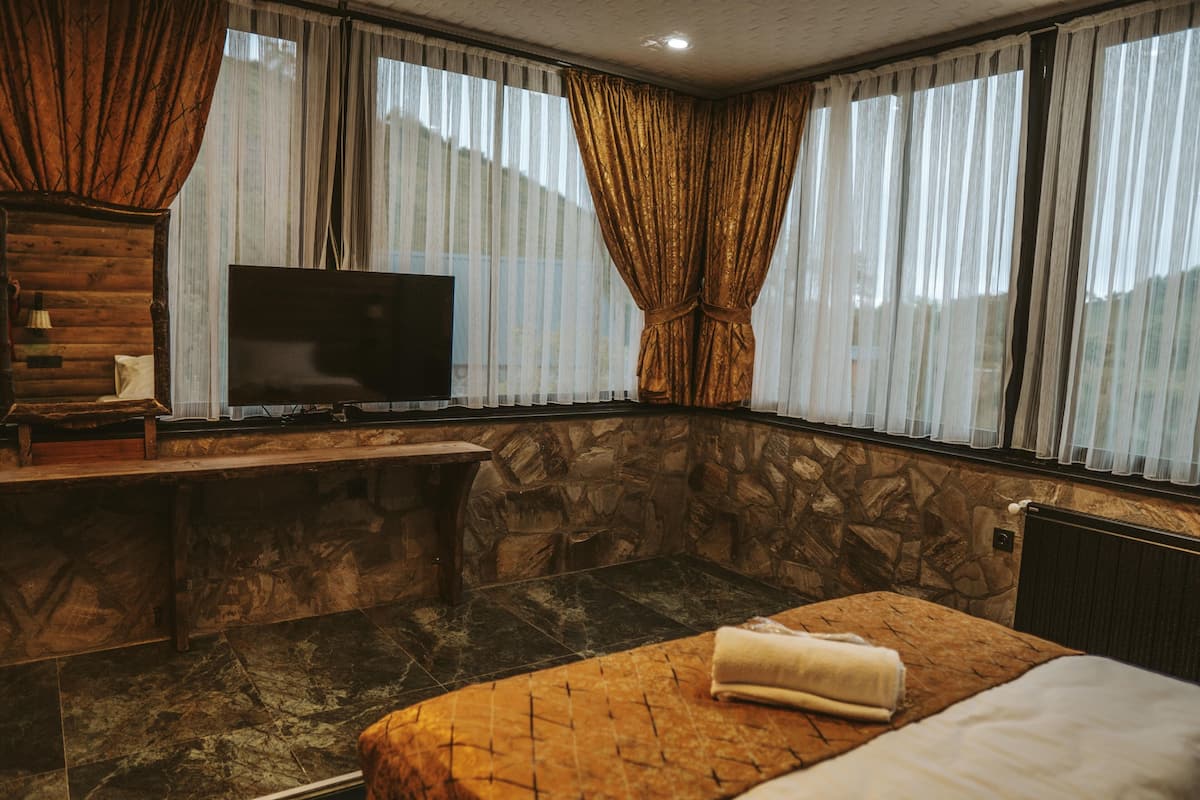 A room with a stone wall, a bed with a brown headboard, a flat-screen TV on a wooden console, and a window with sheer curtains.