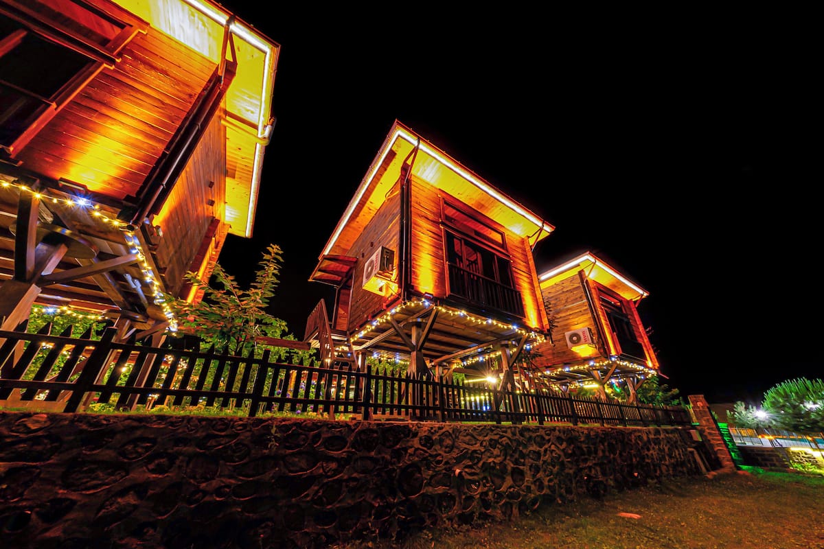 A wooden cabin with a stone foundation, illuminated by warm lights, and surrounded by a wooden fence.