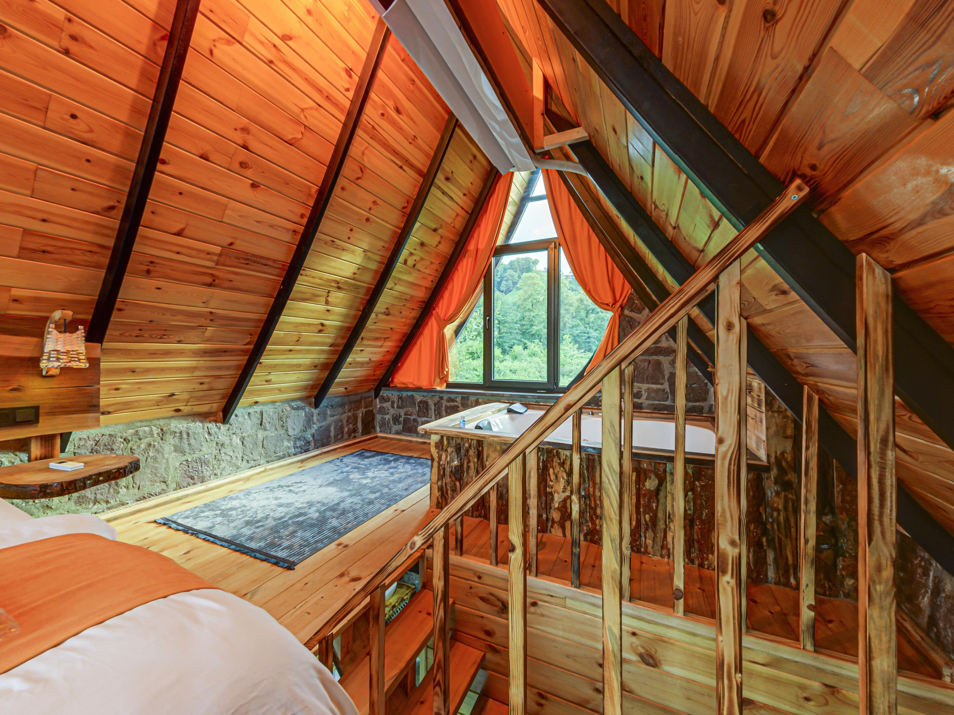 Ein Holzinnenraum mit schrägverdeckter Decke, einer Steinwand, einem Fenster mit Vorhängen und einer Holztreppe.