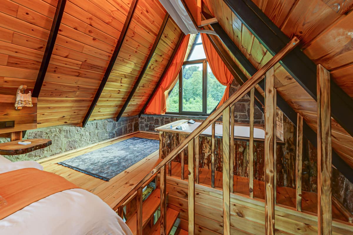 A wooden interior with a sloped ceiling, a stone wall, a window with curtains, and a wooden staircase.