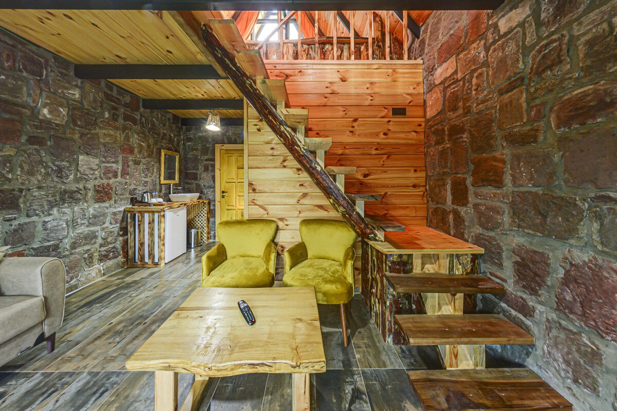Um interior rústico com escada de madeira, paredes de pedra, uma mesa de madeira e duas poltronas amarelas.