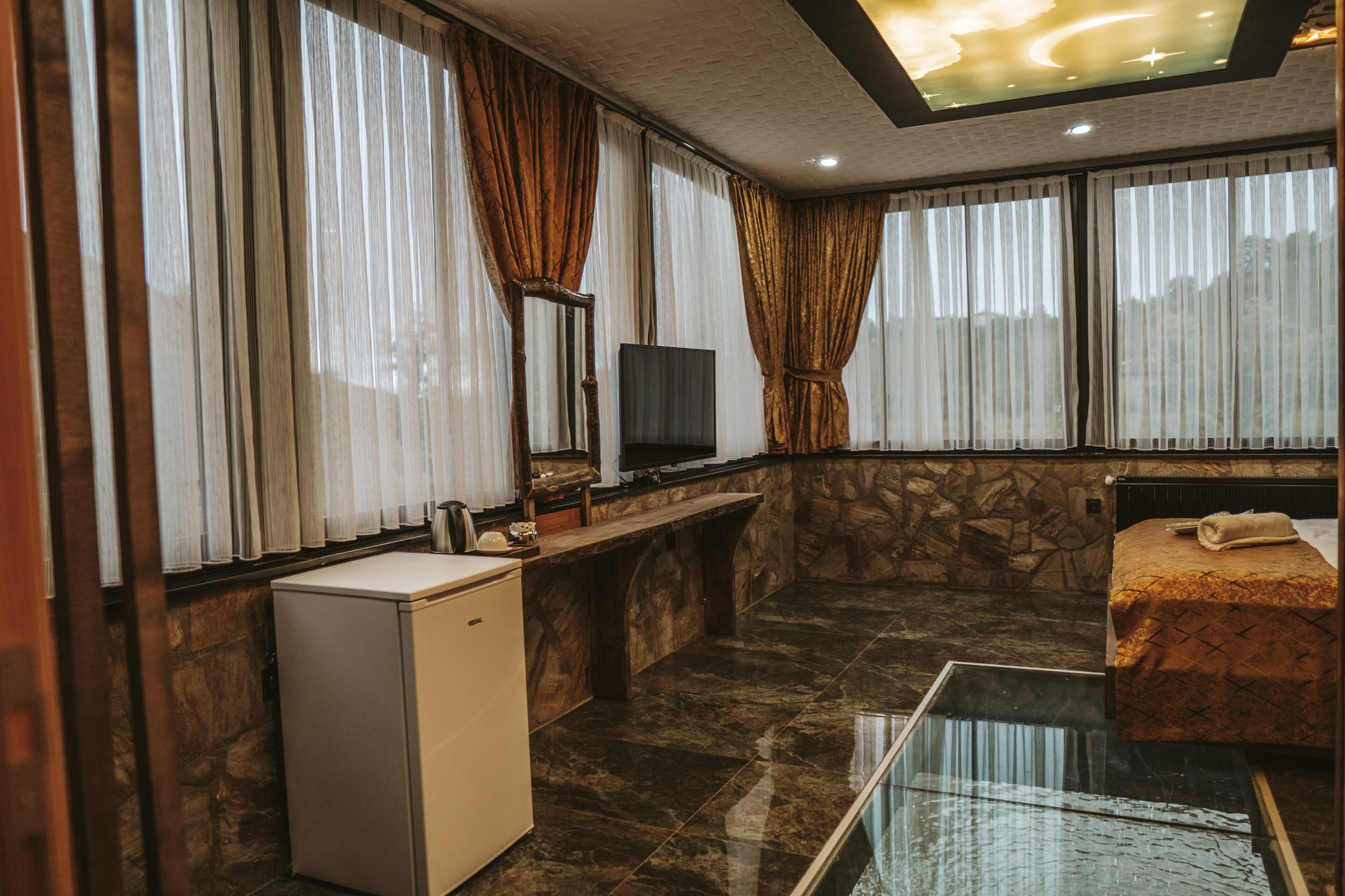 Habitación de hotel con pared de piedra, cama con colcha estampada, refrigerador blanco pequeño, televisor y una mesita de vidrio.
