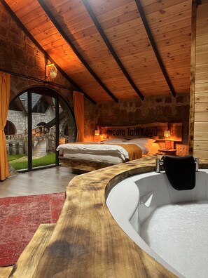 A rustic bedroom with a stone wall, wooden ceiling beams, and a large window. There is a bed, a bathtub, and a small table with a lamp.