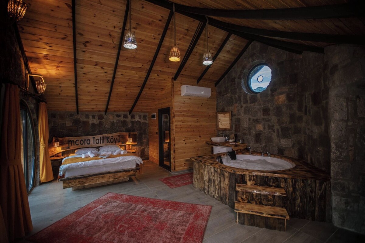 Quarto rústico com teto de madeira, parede de pedra, janela redonda, cama, banco de madeira e pia de pedra.