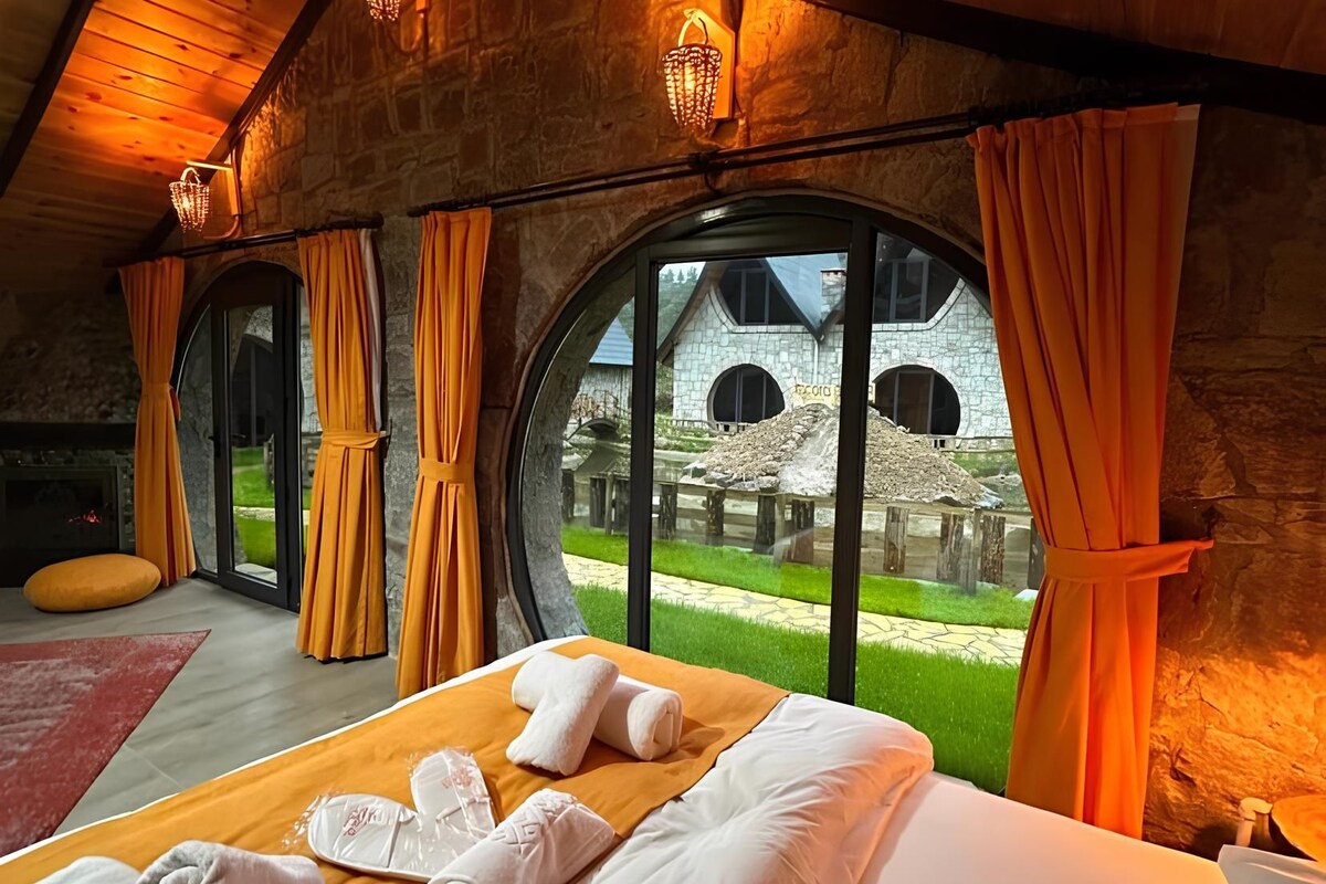 A bedroom with a large bed, a fireplace, and a view of a stone building through large windows.